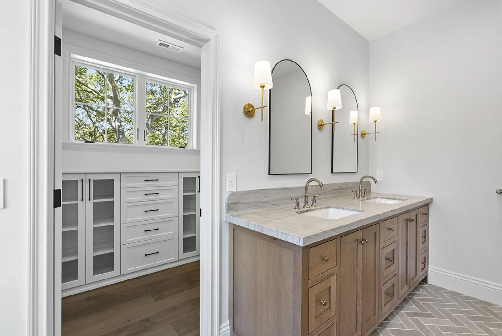 Double-sink oak vanity with stone counter, arched mirrors and brass sconces, walk-in closet beyond